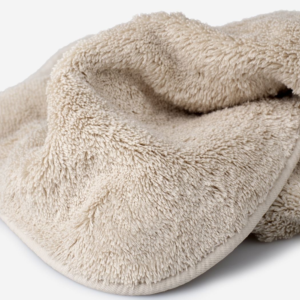 Close-up of a beige cotton towel highlighting the soft terry texture, rounded corner finish, and subtle embroidered logo detail.