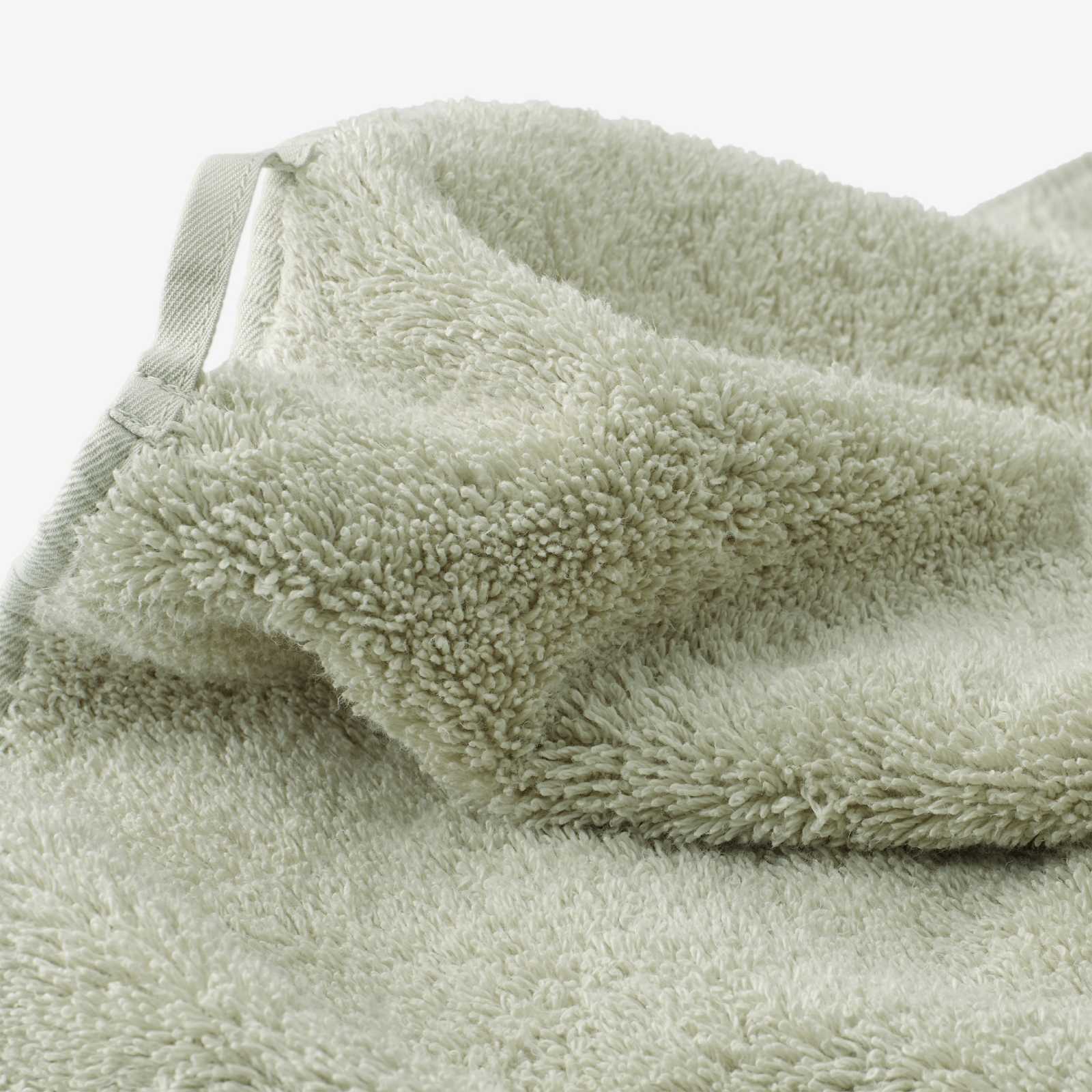 Close-up of a sage green cotton towel showcasing the dense terry texture, folded edge, and integrated fabric hanging loop on a light background.