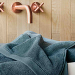 Teal towel draped over a bathtub with copper faucet and handles against a wooden wall.