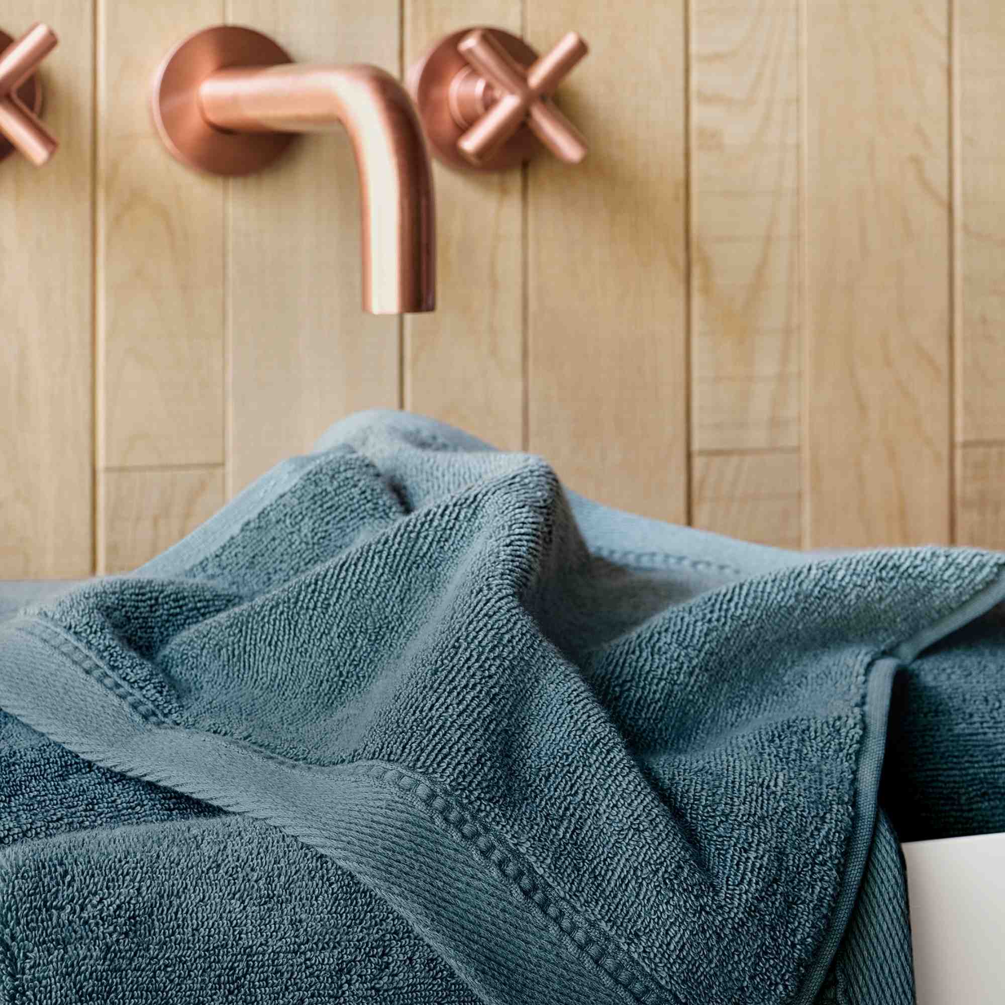 Teal towel draped over a bathtub with copper faucet and handles against a wooden wall.